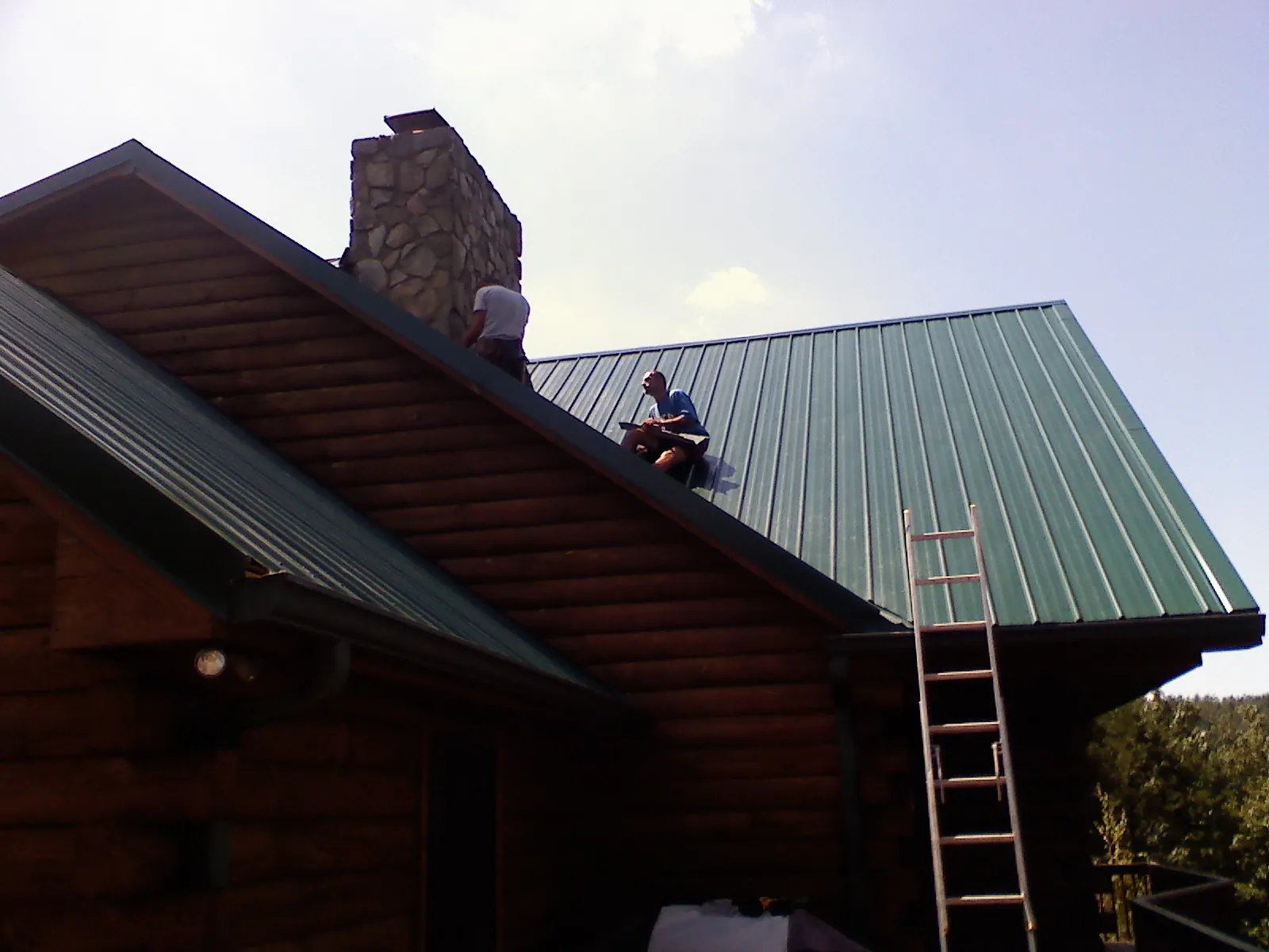Roofers installing green metal panels beside a stone chimney on a log home in Russellville